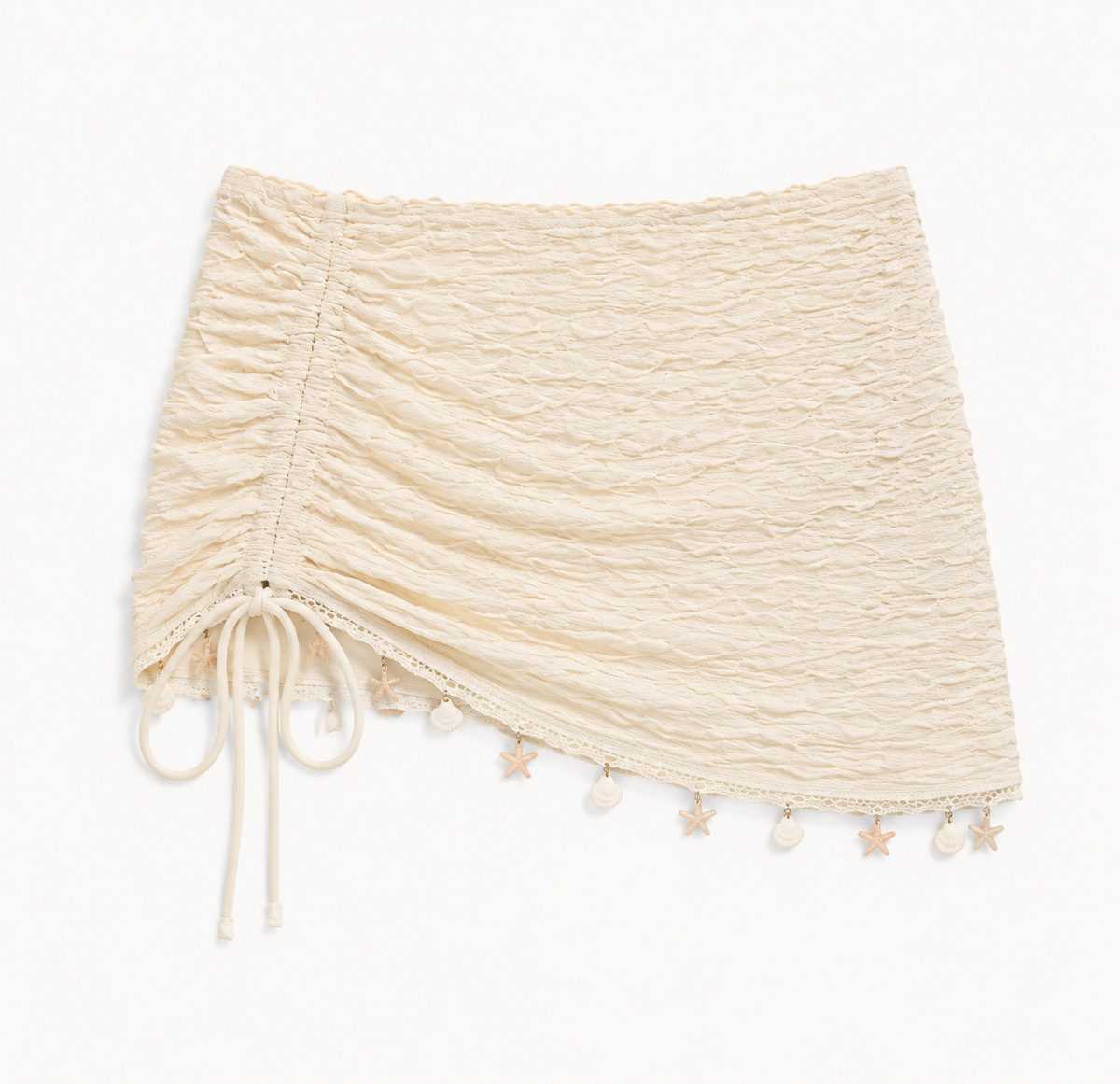 Cream ruched mini cover-up detail showing the side tie, diagonal trim, and shell-like hem accents.