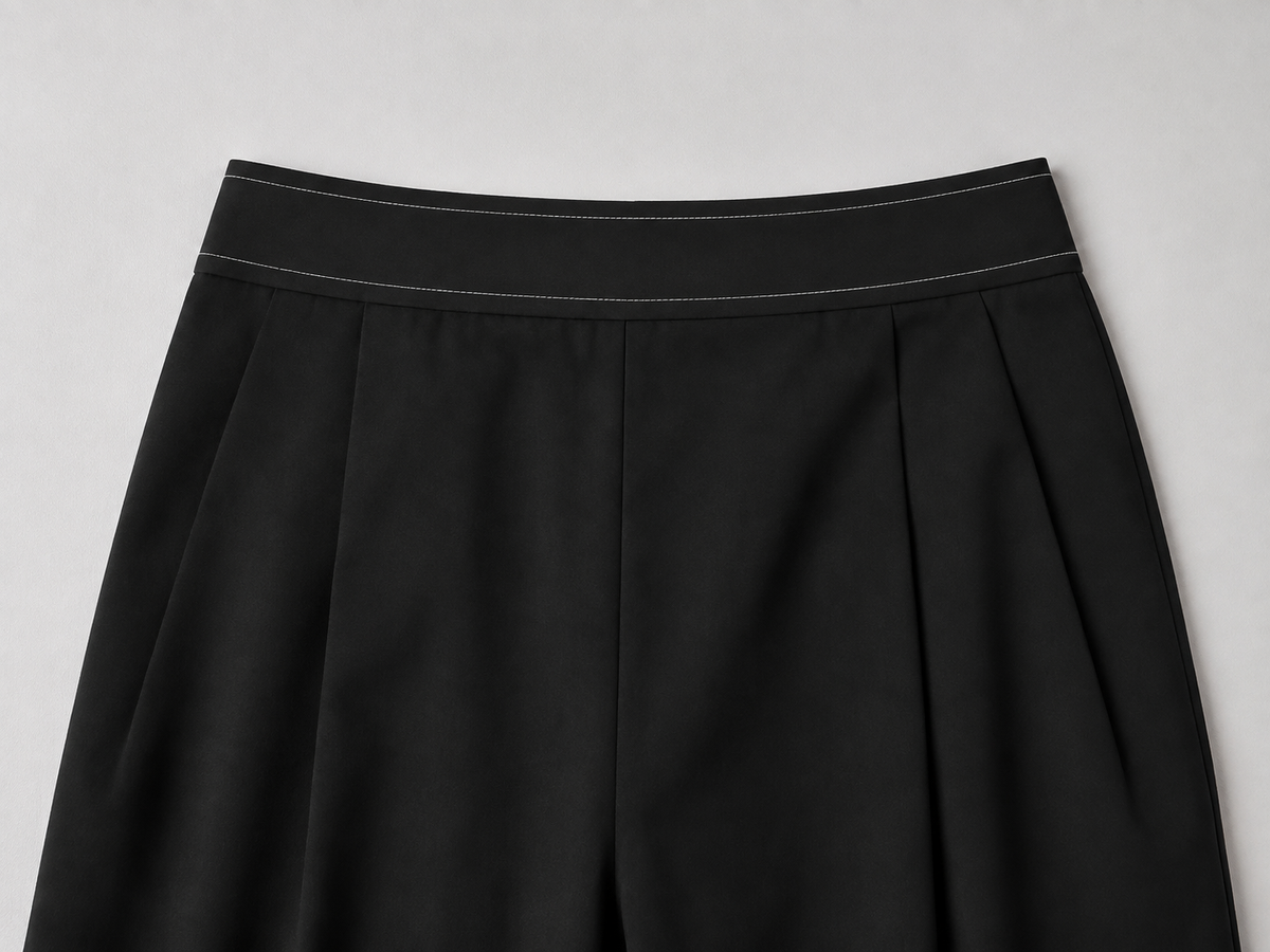 Black wide-leg pants detail showing front pleats and fine contrast waistband lines.